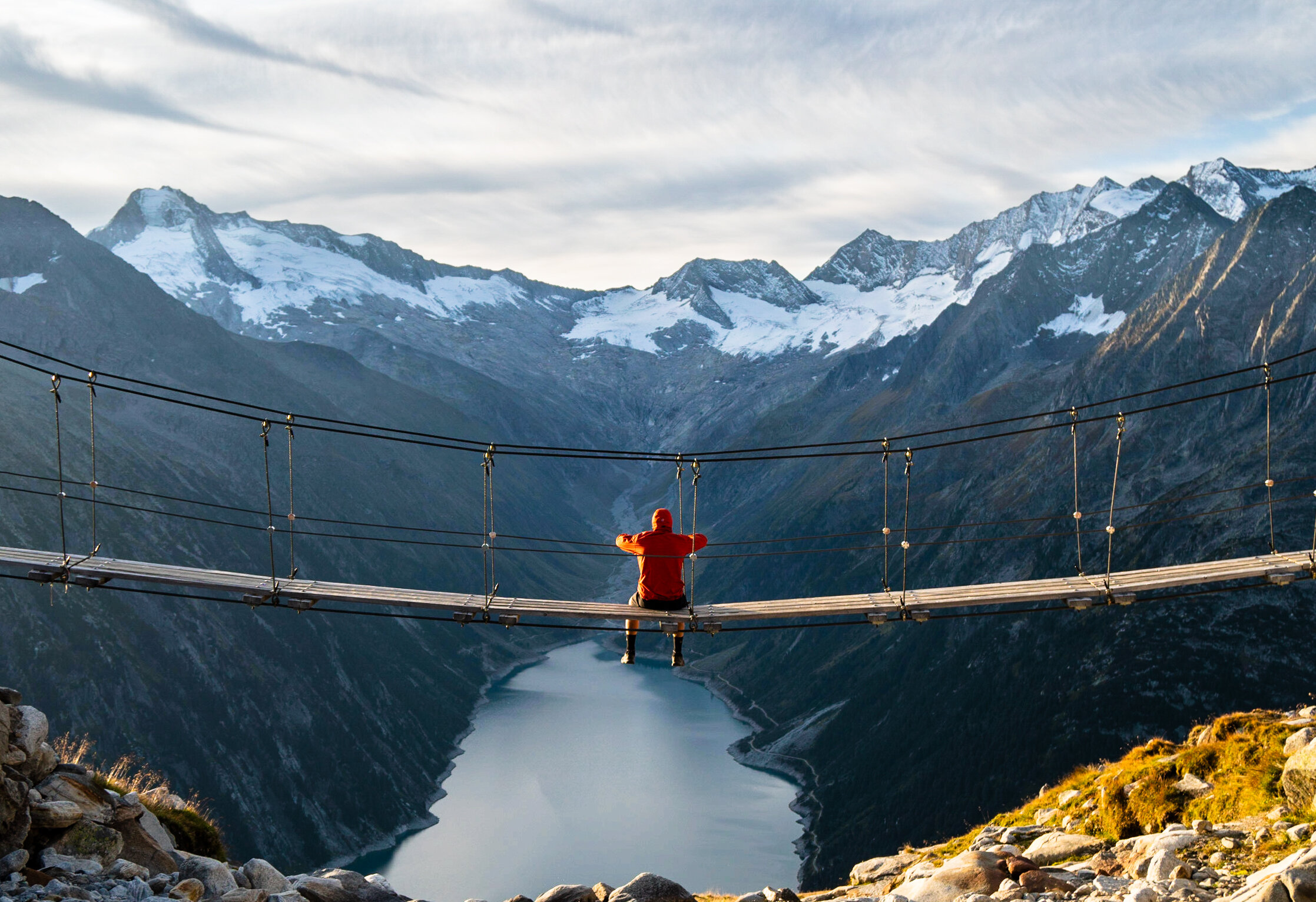 A person in a bright orange jacket sits contemplatively on a suspension bridge spanning a dramatic alpine valley, with snow-capped mountains and a serene lake below, symbolizing the connection and bridging of opportunities